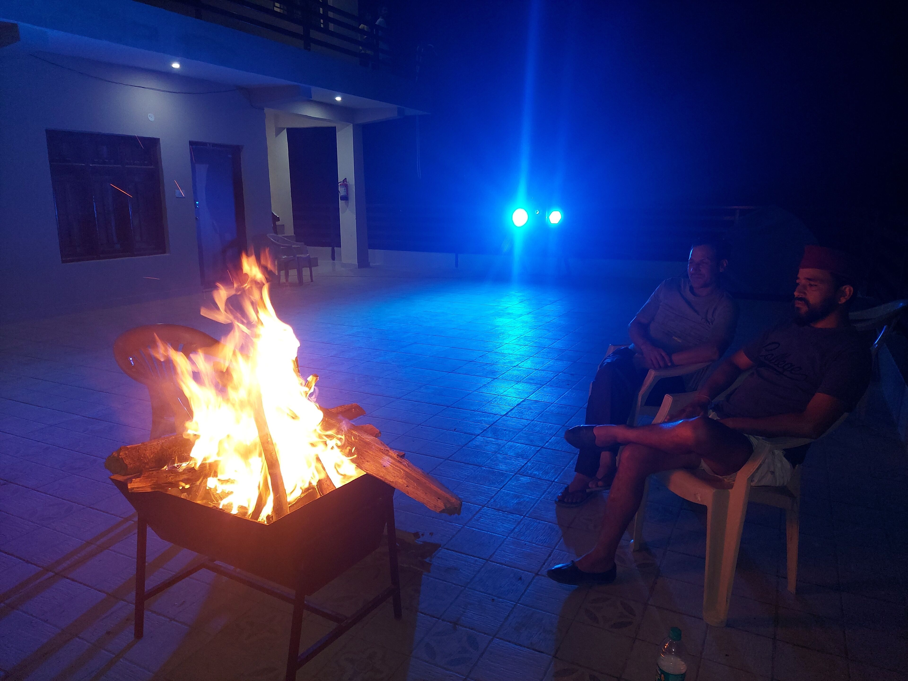 Two people sitting by a fire pit in an outdoor area with tiled flooring and a building in the background.