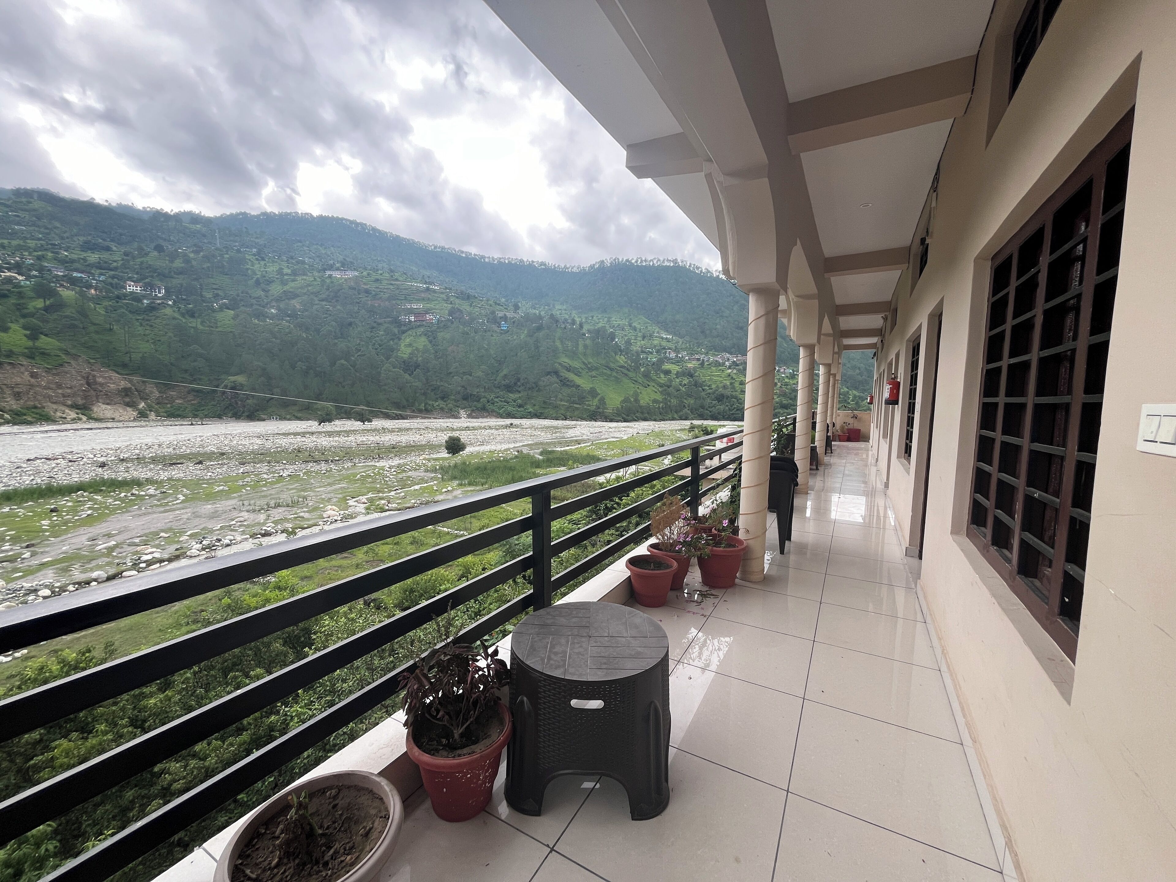 A balcony with potted plants, a small stool, and a view of a river and mountains.
