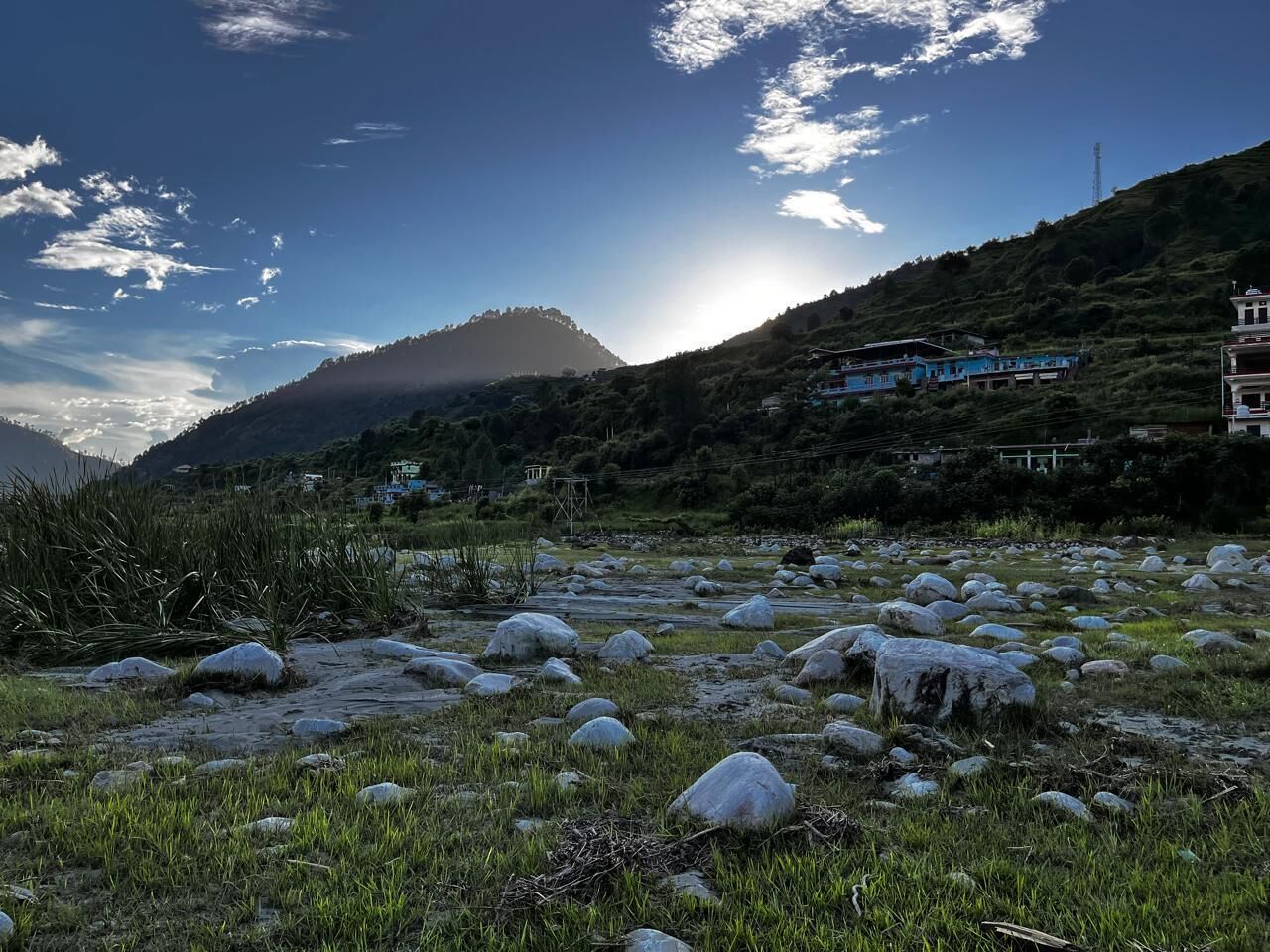 Un paisaje rural con rocas dispersas y hierba, una colina al fondo y un edificio con techo azul.