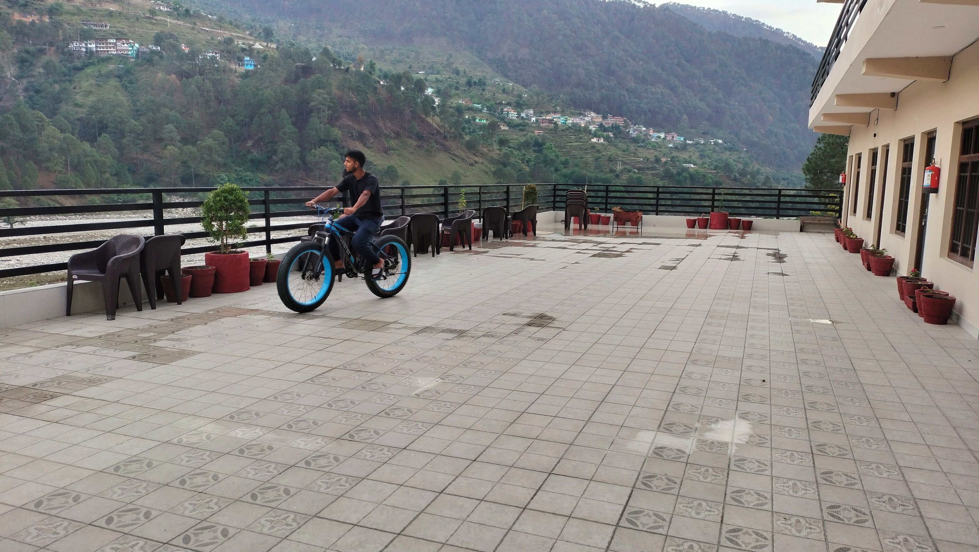 A man riding a blue electric bike on a tiled terrace with potted plants and chairs.