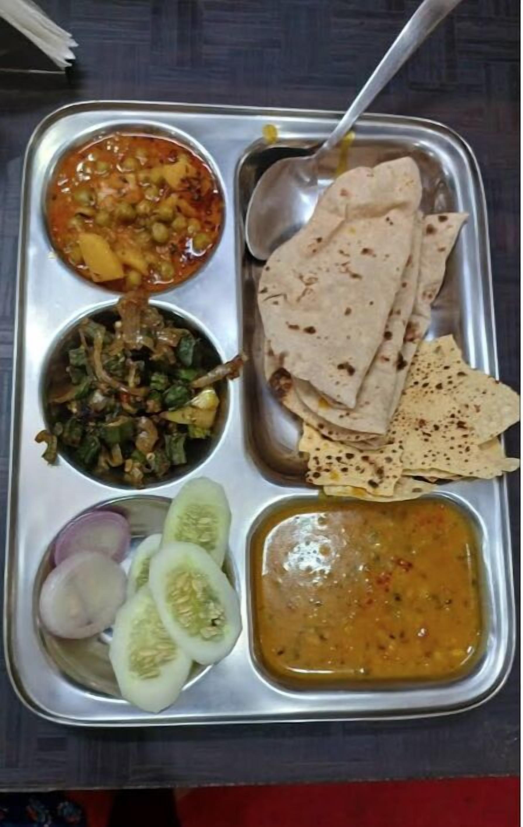 A stainless steel tray with various dishes including a vegetable curry, a lentil dish, a spinach dish, a cucumber salad, and a lentil soup.