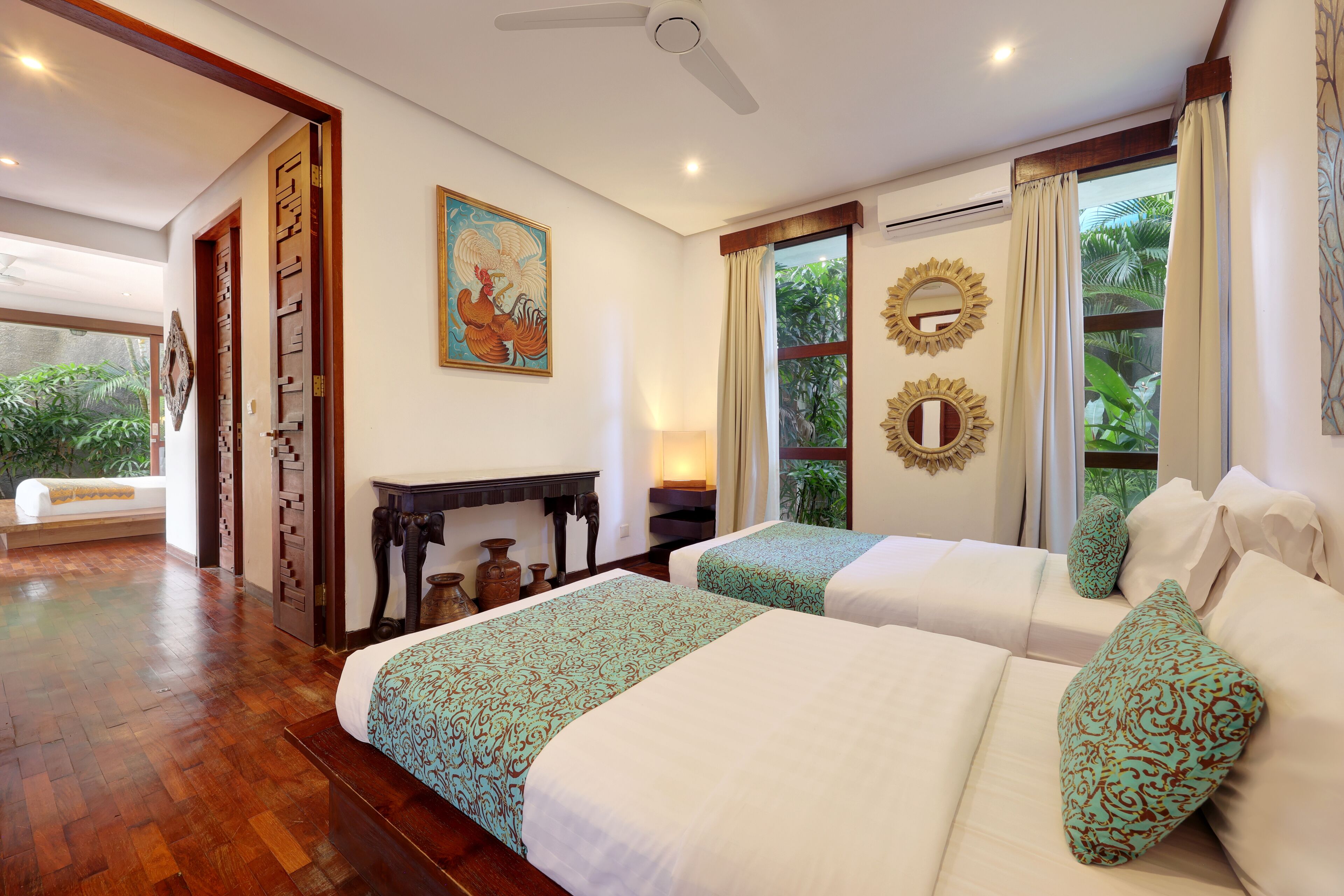 A hotel room with two beds, wooden flooring, a ceiling fan, a painting, and a view of greenery through the window.