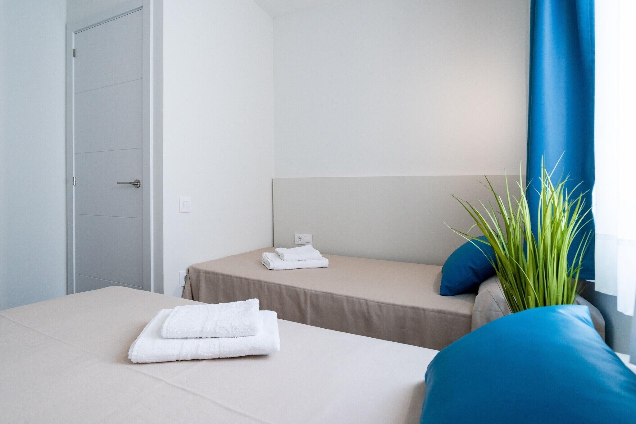 A hotel room with two beds, a headboard, a potted plant, and a window with blue curtains.