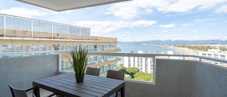 A balcony with a table and chairs, overlooking a coastal cityscape with buildings and a beach.
