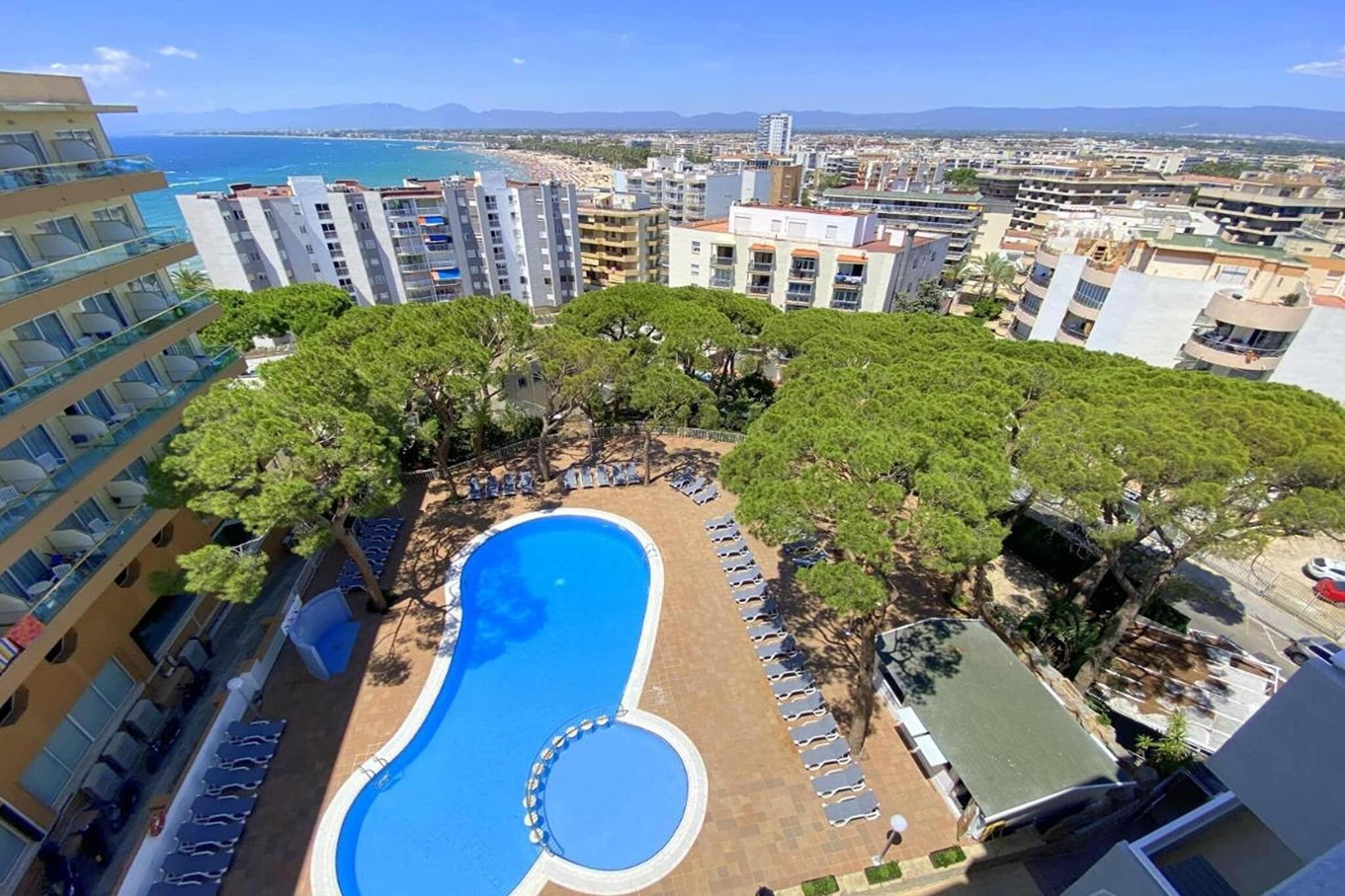 A rooftop pool surrounded by trees and lounge chairs, with a view of the sea and cityscape.