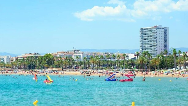 A beach with clear blue water, palm trees, and a tall hotel building in the background. - Chic Stay by the Beach (Tarragona)