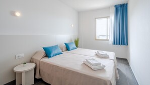 A hotel room with two beds, a small round table, a potted plant, and a window with blue curtains. - Chic Stay by the Beach (Tarragona)