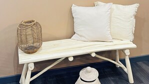 Una habitación con un reloj de pared, un banco blanco con cojines, una cesta tejida y un sombrero tejido en el suelo.