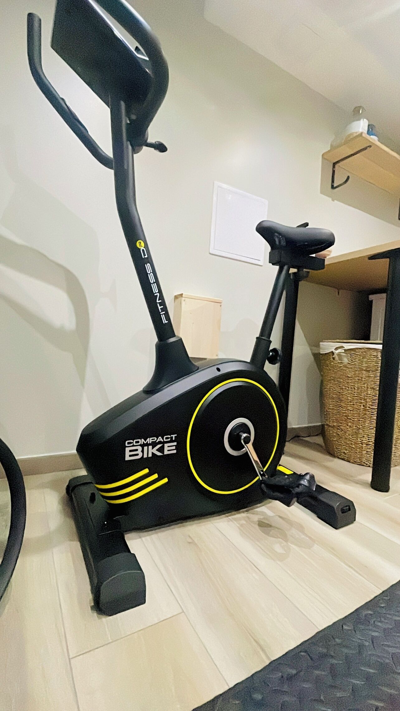 A compact bike exercise machine in a room with a tiled floor, a wooden cabinet, and a wicker basket.