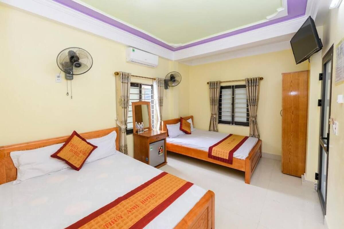 Habitación de hotel con tres camas individuales, cada una con cabecero de madera y colcha a rayas rojas y blancas. Hay dos ventiladores de pared, un armario de madera y un televisor montado en la pared.