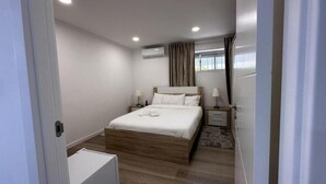 Un dormitorio moderno con una cama de madera, ropa de cama blanca, dos mesitas de noche con lámparas, una ventana con cortinas y un equipo de aire acondicionado.