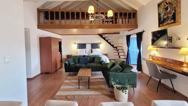 A spacious living area with a high ceiling, a wooden staircase, a green sofa set, a dining table with chairs, and a large window.