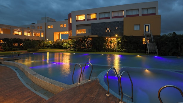 A swimming pool with a wooden deck and metal railings, surrounded by lush greenery and a modern building with illuminated windows. - Sinclairs Burdwan (Burdwan)