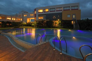 A swimming pool with a wooden deck and metal railings, surrounded by lush greenery and a modern building with illuminated windows. - Sinclairs Burdwan (Burdwan)