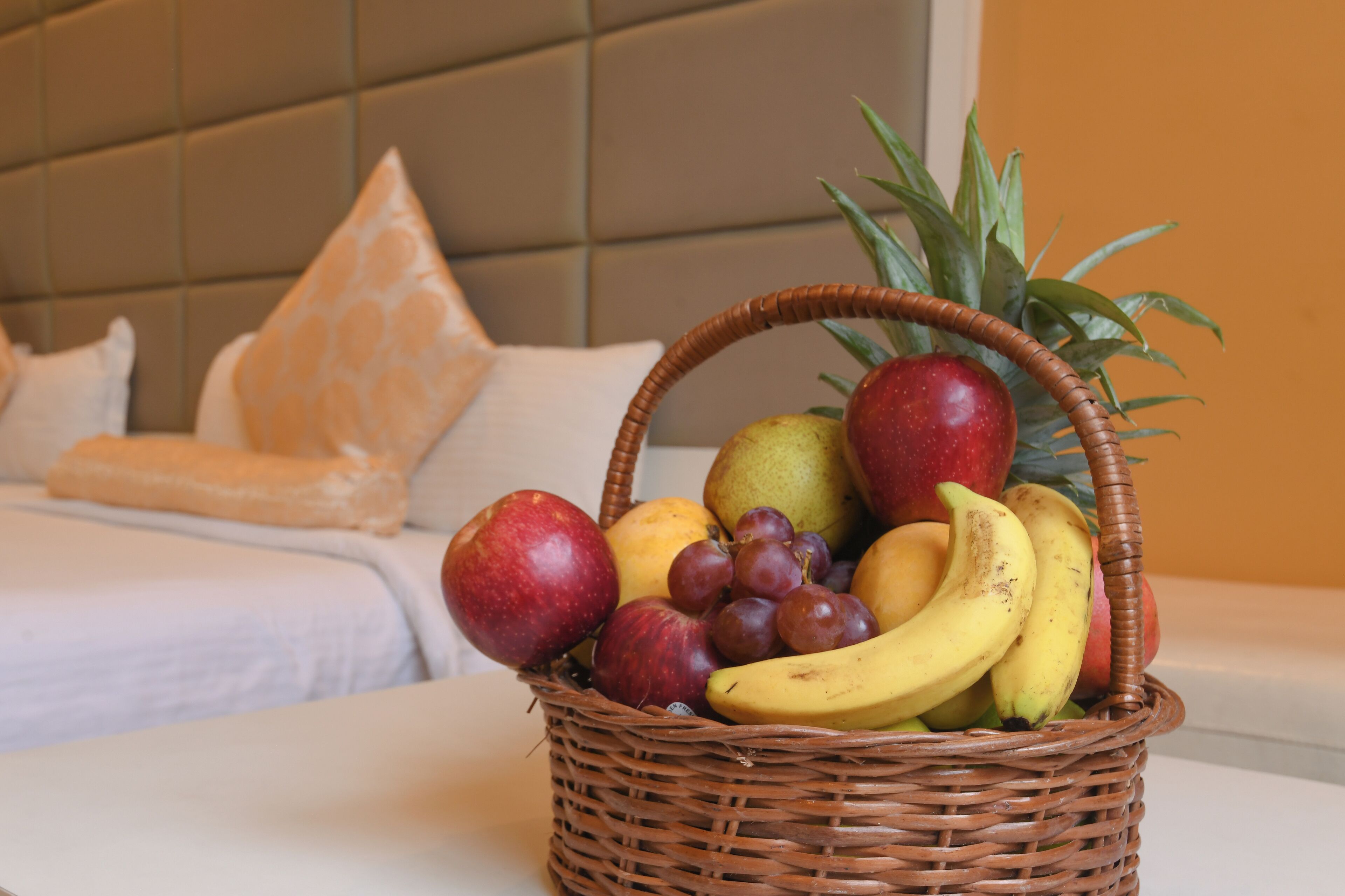 Una cesta de mimbre con fruta sobre una mesa, con una cama y almohadas al fondo.