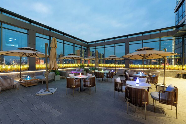 An outdoor seating area with tables and chairs, umbrellas, and a view of the cityscape through large windows.
