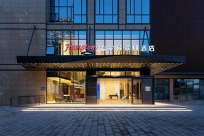 Una entrada de hotel moderna con fachada de vidrio, un llamativo letrero de 'Hampton by Hilton' y un área de acceso cubierta.