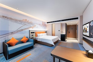 A modern hotel room with a bed, a sofa, a desk, and a large cityscape mural on the wall.