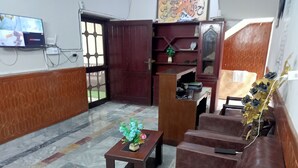 A lobby with a brown leather sofa, a small wooden table with a floral arrangement, a TV mounted on the wall, and a patterned floor. - Welcome Inn Guest House (Islamabad)