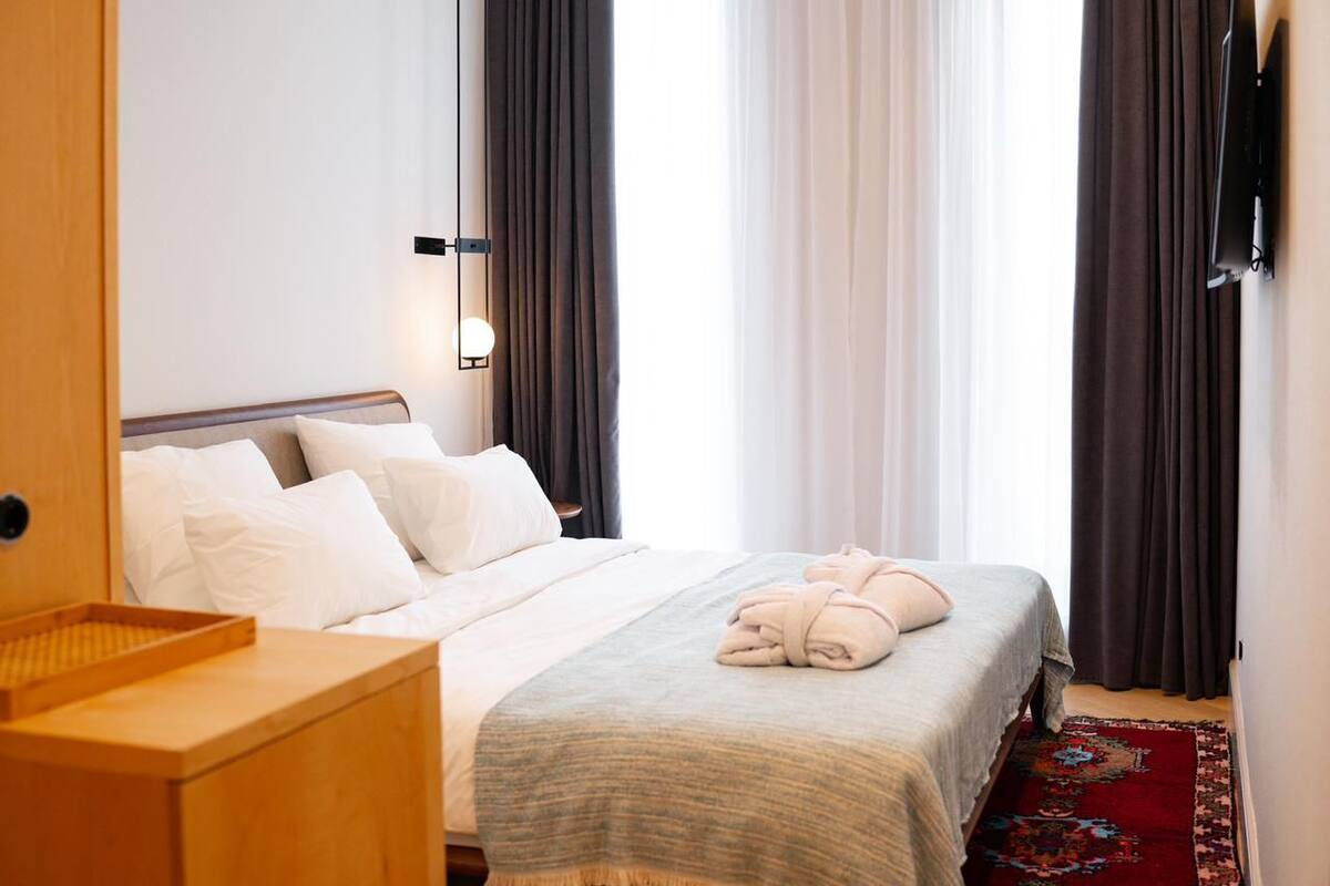 Une chambre d’hôtel avec un lit, une table de chevet en bois, un téléviseur fixé au mur et une fenêtre avec des rideaux.