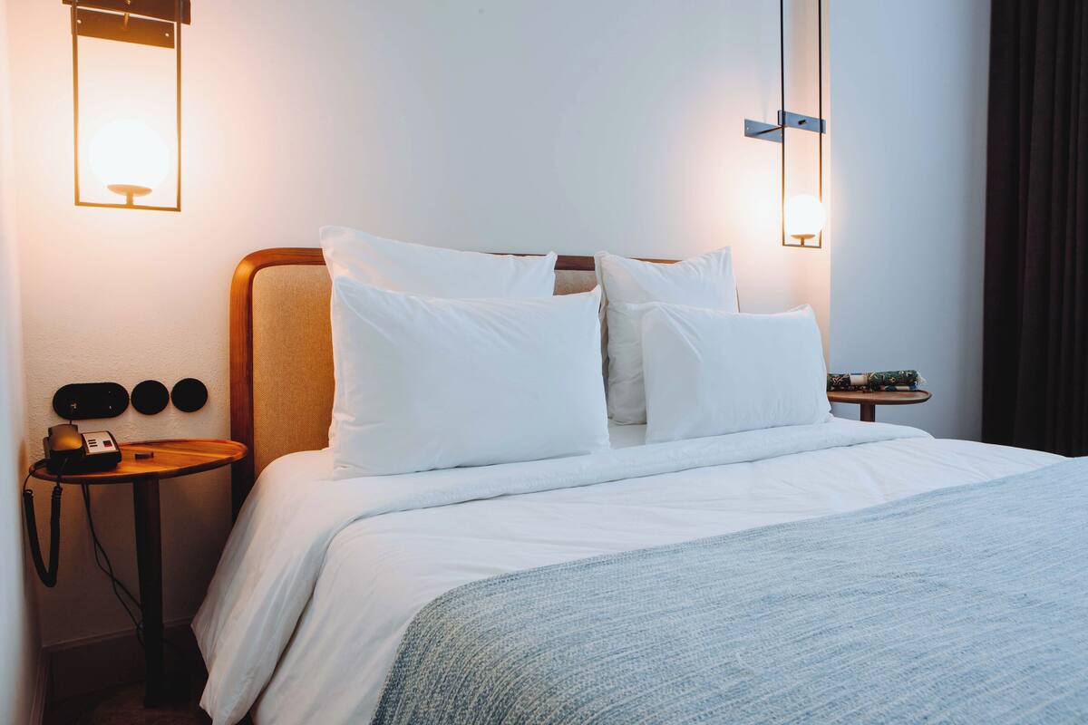 Un lit bien fait, avec une literie blanche et un plaid bleu, une table de chevet avec une lampe et un luminaire fixé au mur.