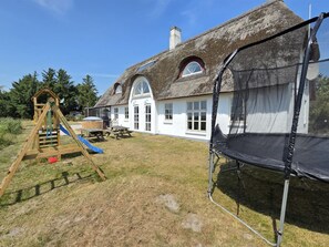 Une maison à toit de chaume avec une structure de jeu en bois et un trampoline dans la cour.