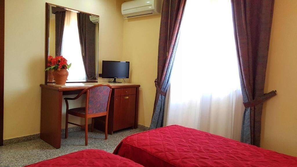 A hotel room with two single beds, a desk with a computer, a chair, a mirror, and a window with curtains.