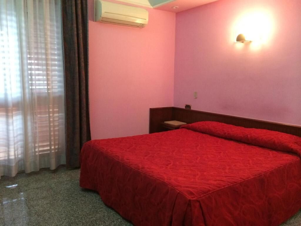 A room with a bed, a nightstand, a wall-mounted light, and a window with curtains.