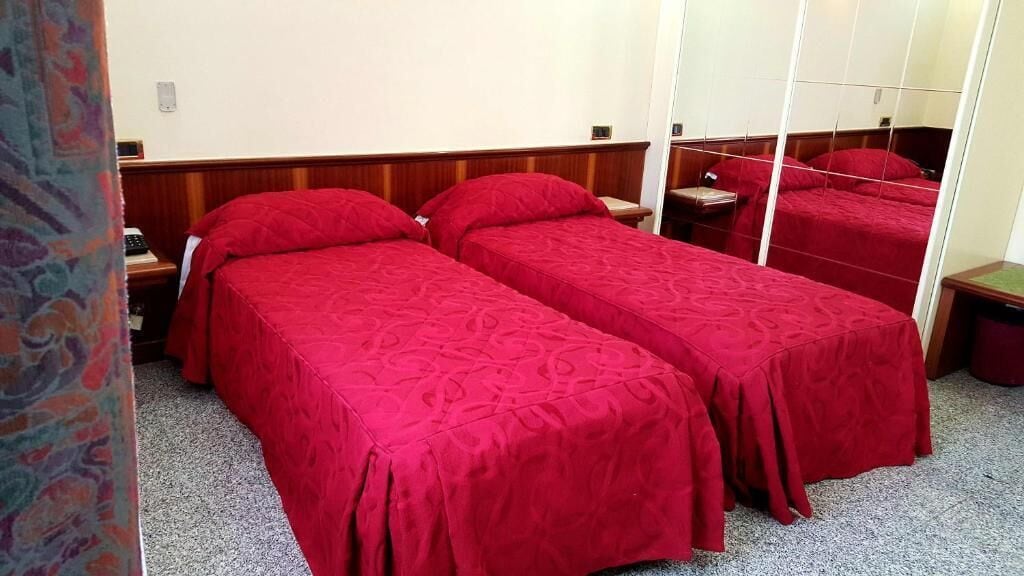 Une chambre d’hôtel avec deux lits simples, chacun recouvert d’une couverture rouge et agrémenté d’oreillers assortis. On y trouve une tête de lit en bois, une table de chevet avec une lampe et un miroir au mur.