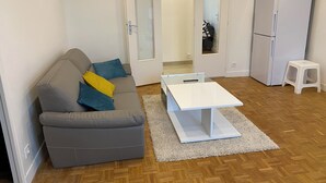 Ein Wohnzimmer mit grauem Sofa, einem weißen Couchtisch, einem weißen Kühlschrank und einem kleinen weißen Hocker.