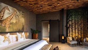 A bedroom with a large bed, giraffe-themed pillows, a wall mural, a fireplace, and a seating area with giraffe-patterned cushions. - Ximuwu Safari Lodge (Klaserie)