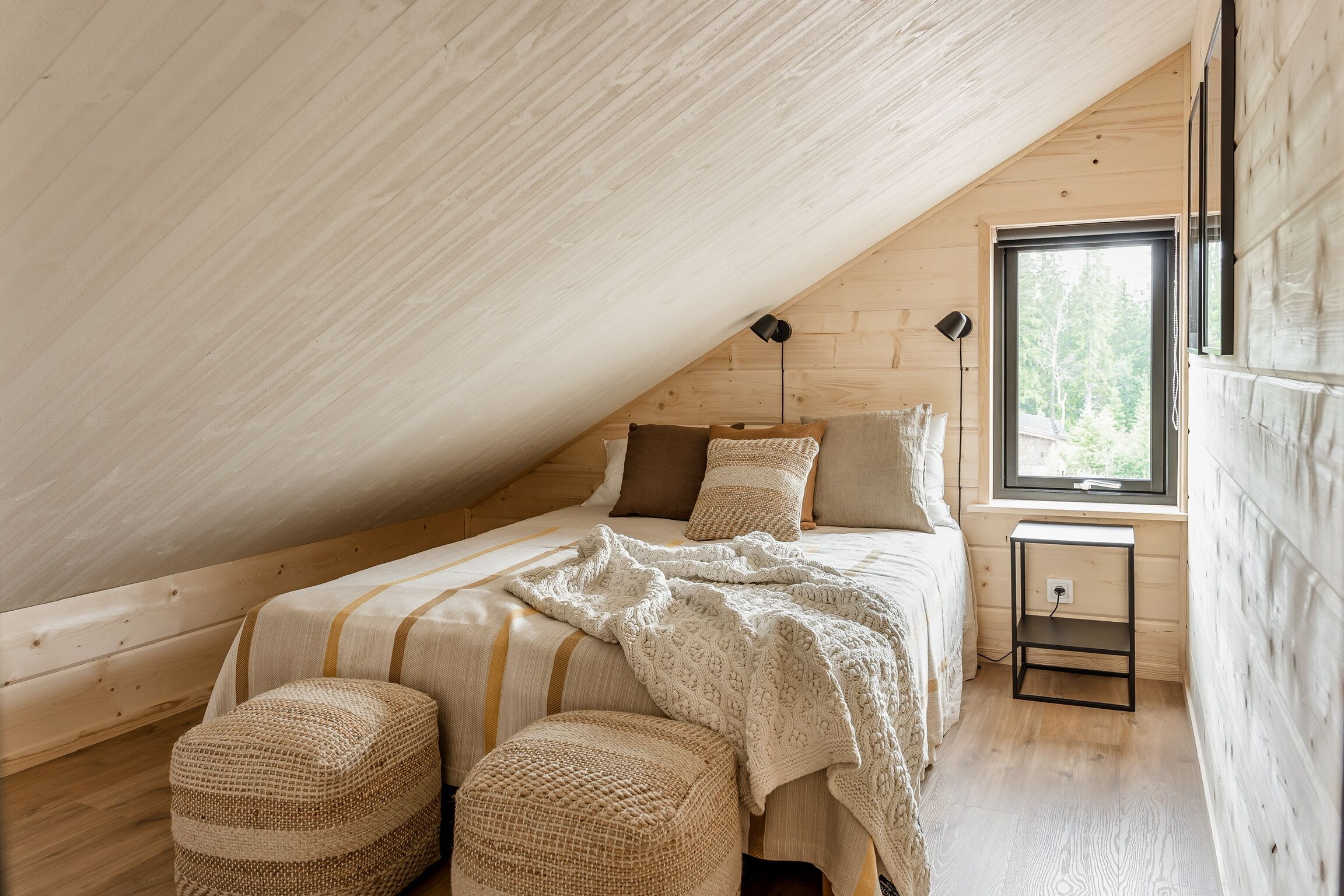 A cozy attic bedroom with a sloped ceiling, a bed with striped bedding, two woven ottomans, a bedside table with a lamp, and a window with a view of greenery.
