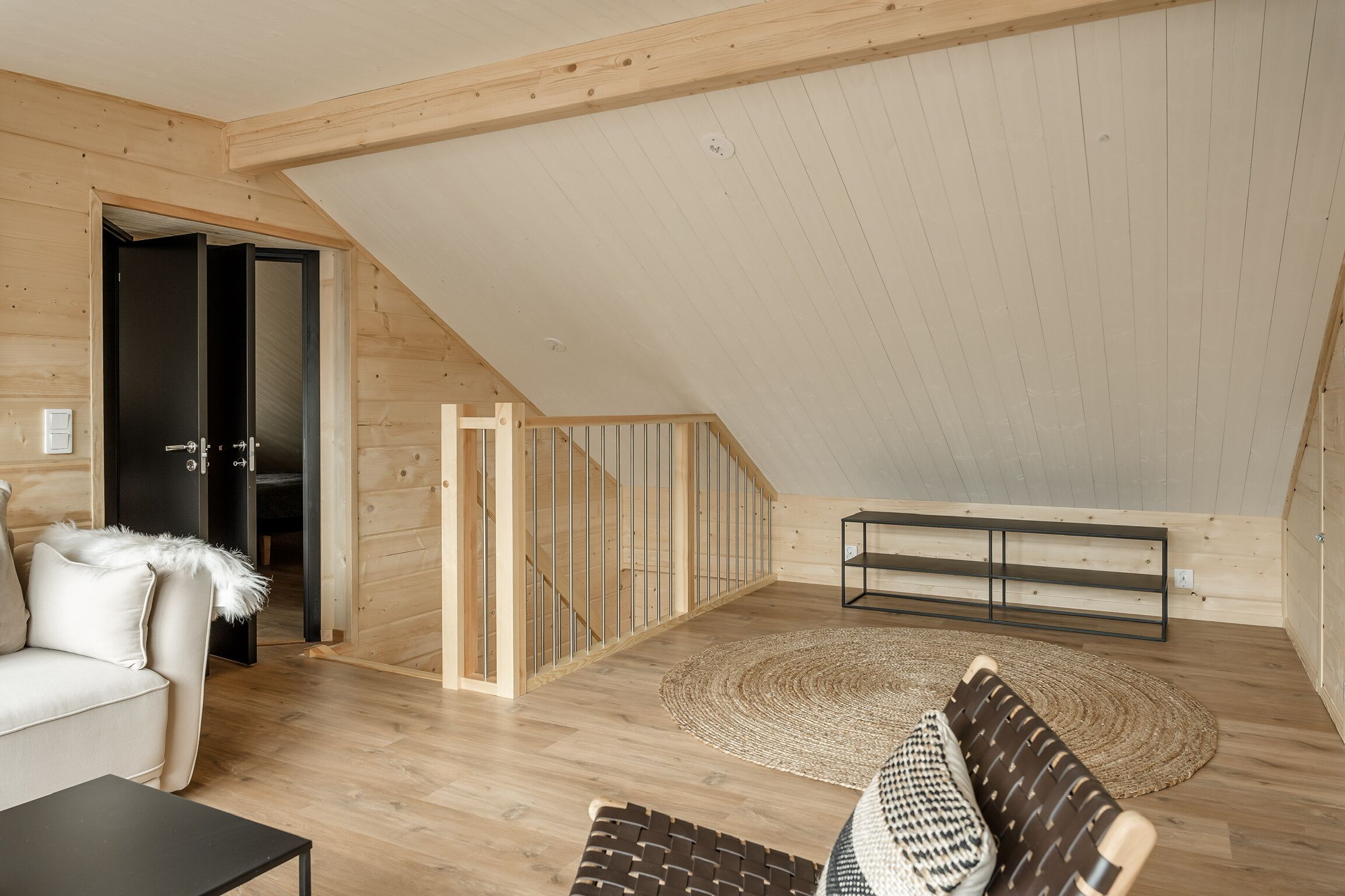 Un'ampia camera da letto mansardata con soffitto spiovente, pavimento in legno, una grande finestra e un'accogliente area salotto con divano e tappeto decorato.
