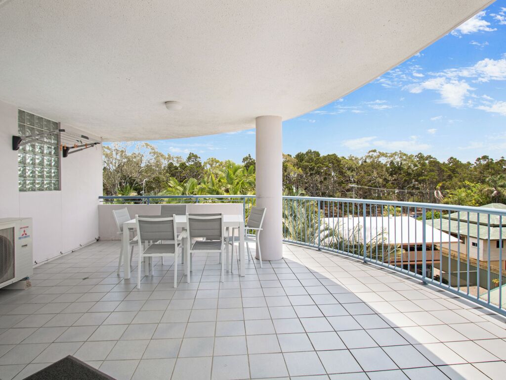 A covered balcony with white chairs and a table, a white air conditioning unit, and a view of greenery and buildings in the background.