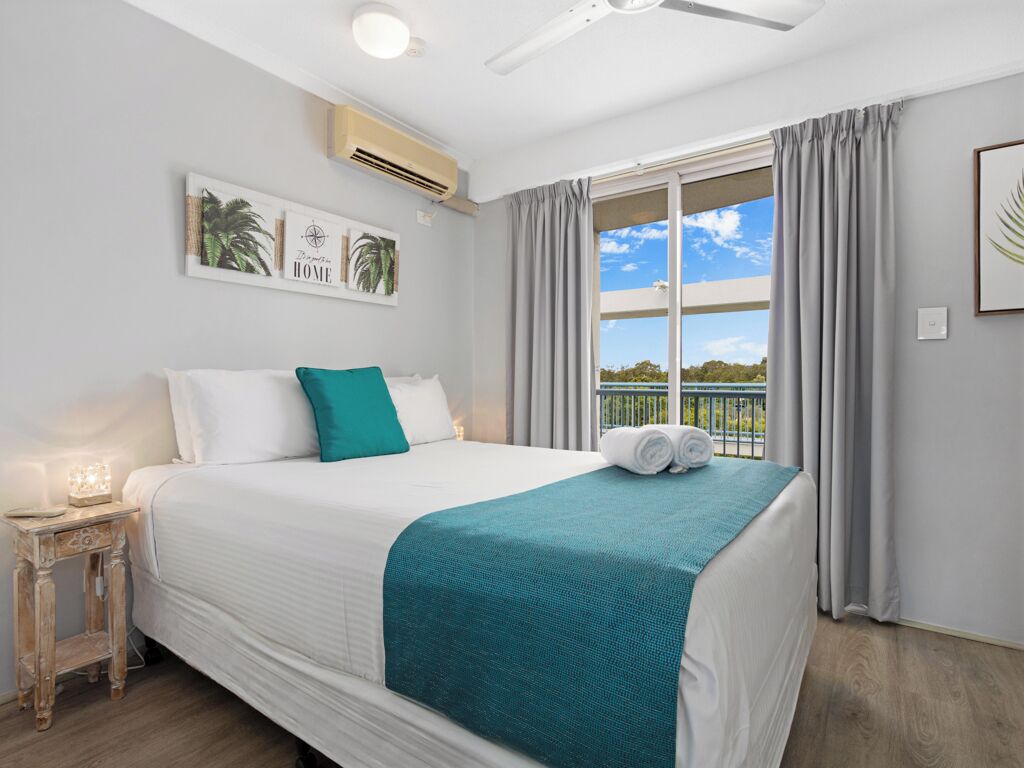 Une chambre d’hôtel avec un grand lit, une table de chevet avec une lampe, un ventilateur de plafond et une fenêtre avec des rideaux.