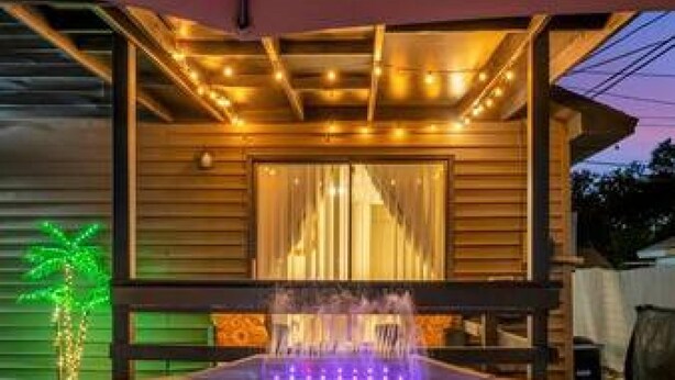 A hot tub with water jets and LED lighting, surrounded by wooden deck and a covered area with string lights. - Family Fun Backyard Oasis Hot Tub Games BBQ (St. Petersburg)