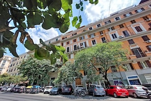A multi-story building with balconies and windows, parked cars, and trees along the street.