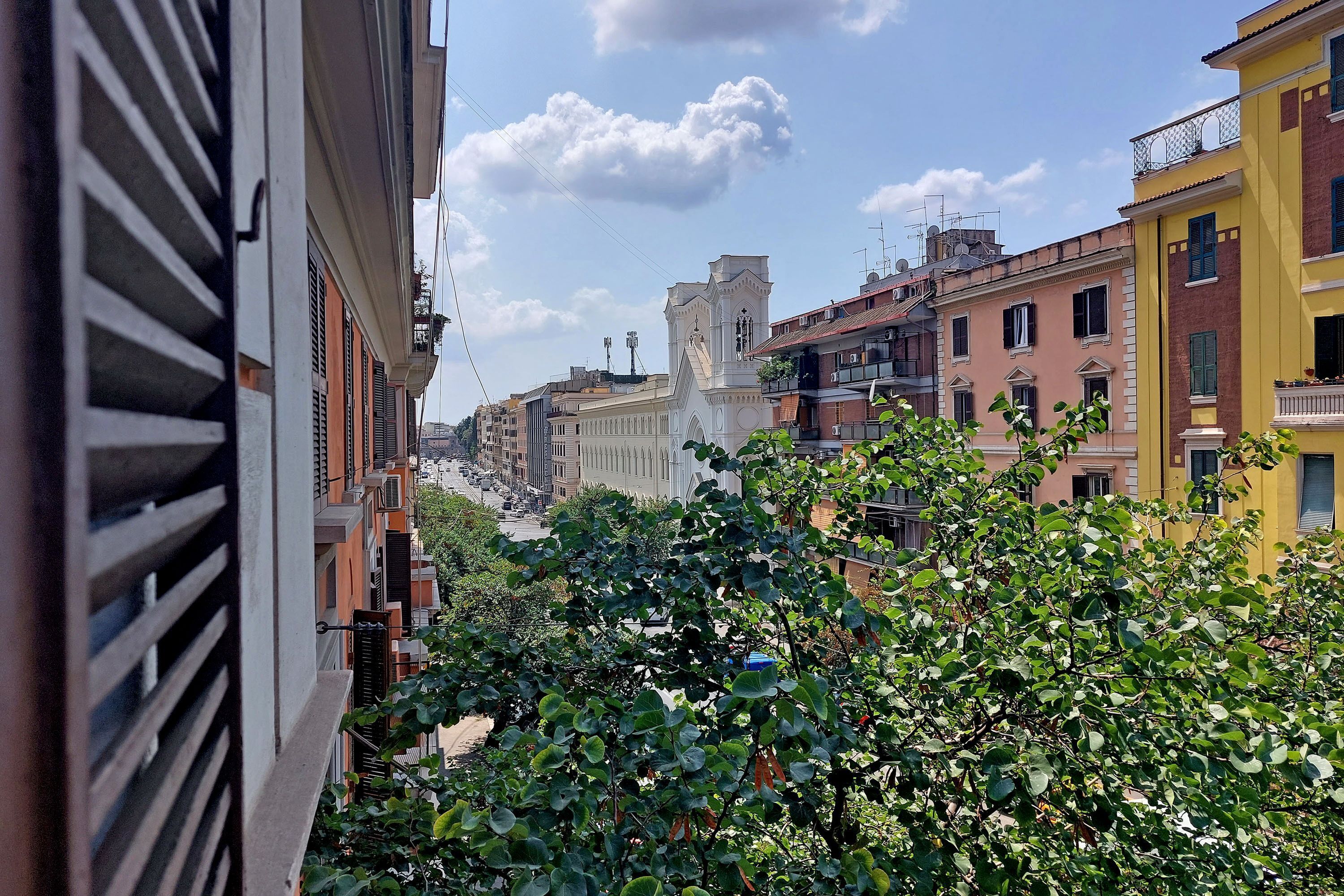 A narrow street with buildings on either side, a prominent white tower, and lush green foliage in the foreground.