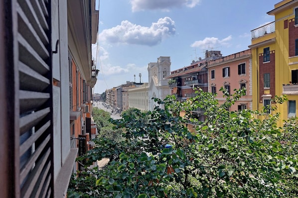 A narrow street with buildings on either side, a prominent white tower, and lush green foliage in the foreground.