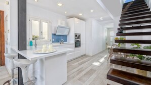 A modern kitchen with white cabinetry, a central island with bar stools, a built-in oven, and a staircase with wooden steps. - Solaga - Noemi Centro (Málaga)