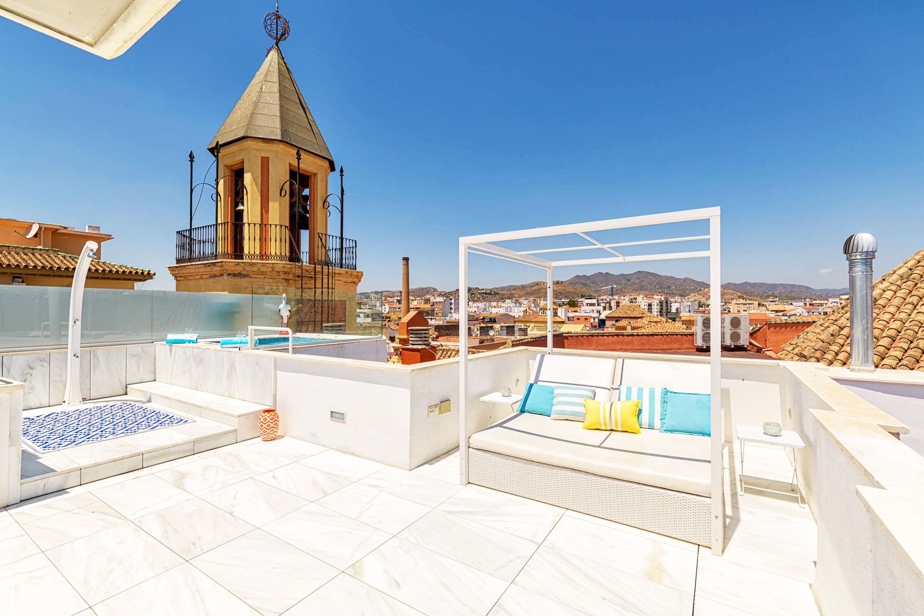 Una terraza en la azotea con una zona de estar exterior blanca, una pequeña estructura tipo torre y una vista despejada del paisaje urbano.
