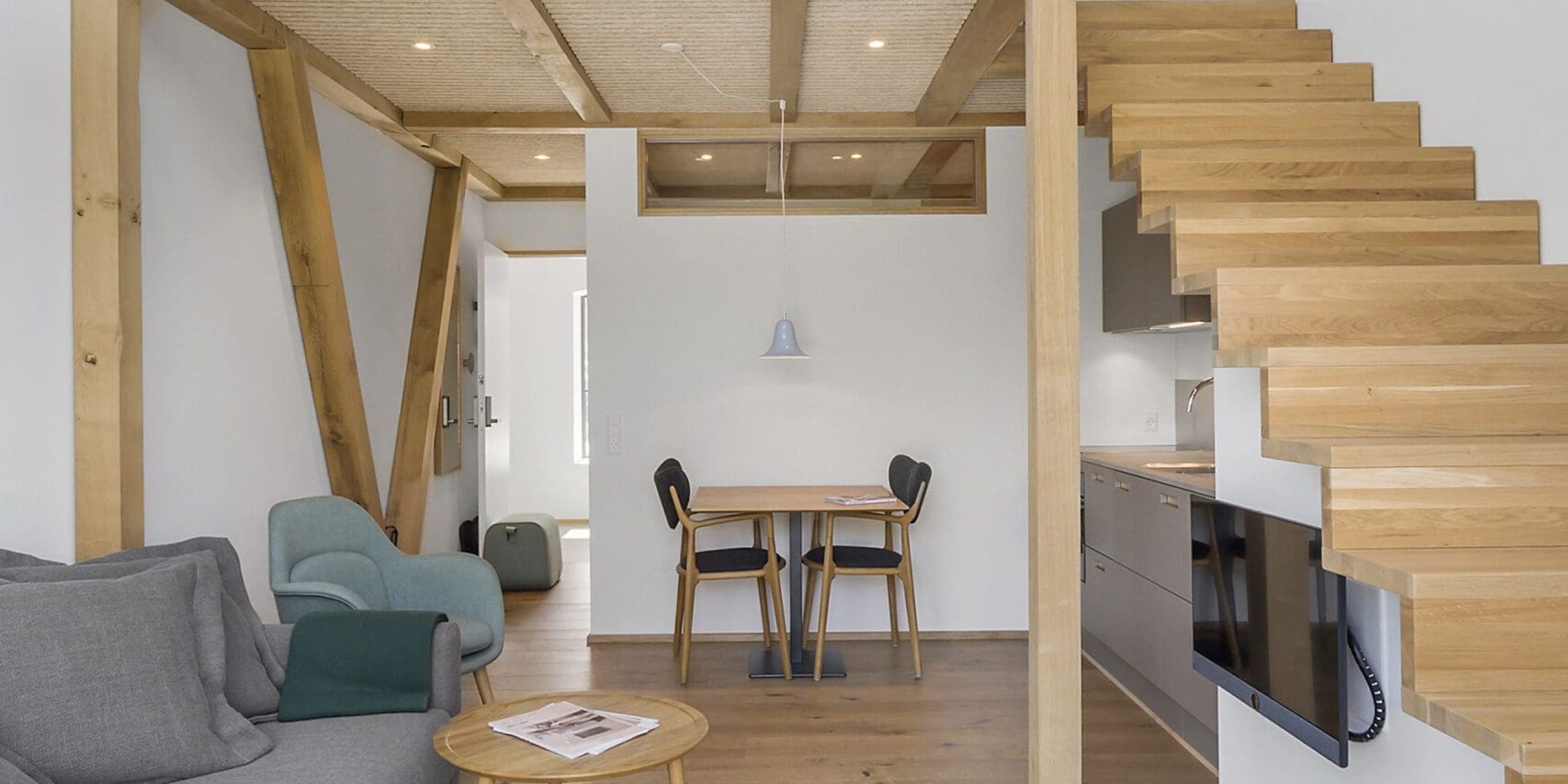Un interior moderno con una escalera de madera, un comedor con dos sillas, un sofá y una mesita con una revista.