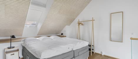 Un dormitorio con techo inclinado, cama con ropa de cama blanca, mesita de noche con lámpara y un espejo grande en la pared.