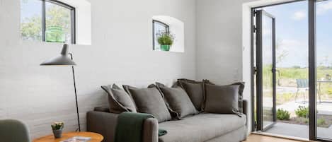 Un salón moderno con un sofá gris, una lámpara de pie, una mesita con una revista y vistas a un espacio exterior por una puerta de cristal.