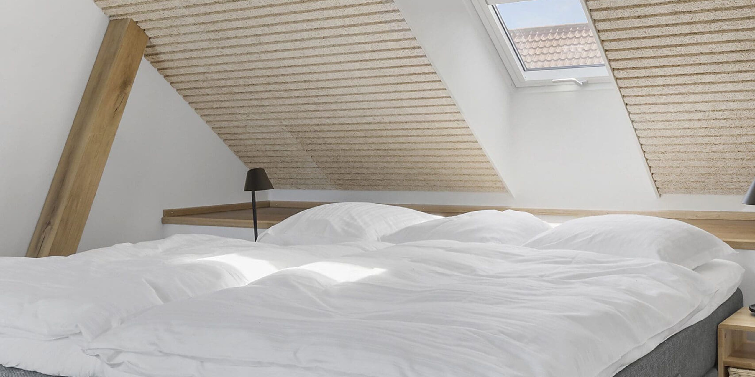 A bedroom with a sloped ceiling, a skylight, a bed with white bedding, and a bedside table with a lamp.