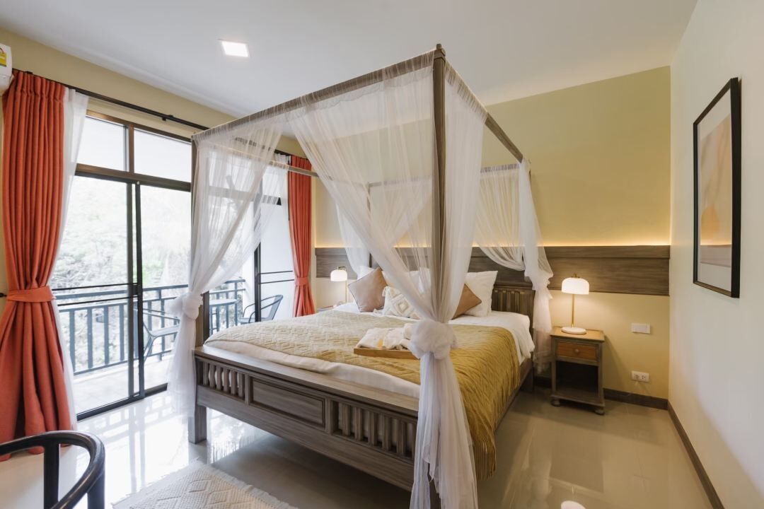 A four-poster bed with a canopy, a bedside table with a lamp, and a view of the outdoors through a large window.