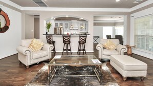 A modern living room with a glass coffee table, two armchairs, and a sofa. A kitchen is visible in the background with bar stools. - REIGN Mini-mansion (Houston)