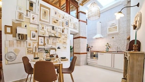 Una cocina moderna con armarios blancos, una zona de comedor con sillas marrones y una pared decorada con cuadros y objetos de arte.