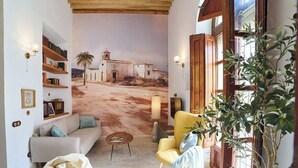 Una sala de estar con un mural de un pueblo, un sofá, un sillón amarillo, un asiento informal y una lámpara, además de una estantería.