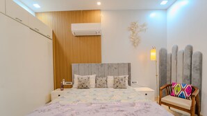 A bedroom with a bed, wooden panel wall, air conditioning unit, decorative wall piece, and a chair with a colorful cushion. - VIBE 2.0 (Jaipur)
